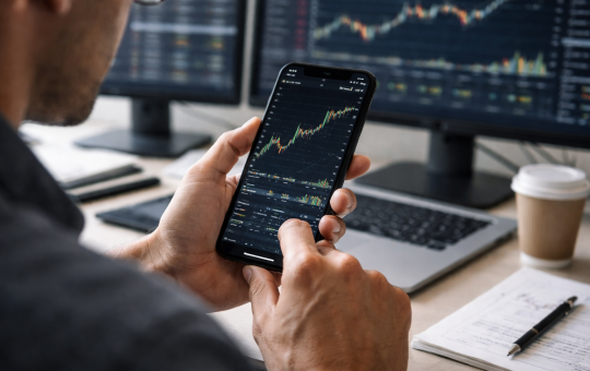A trader analyzes a financial price chart on a smartphone while multiple market charts display on monitors in the background.